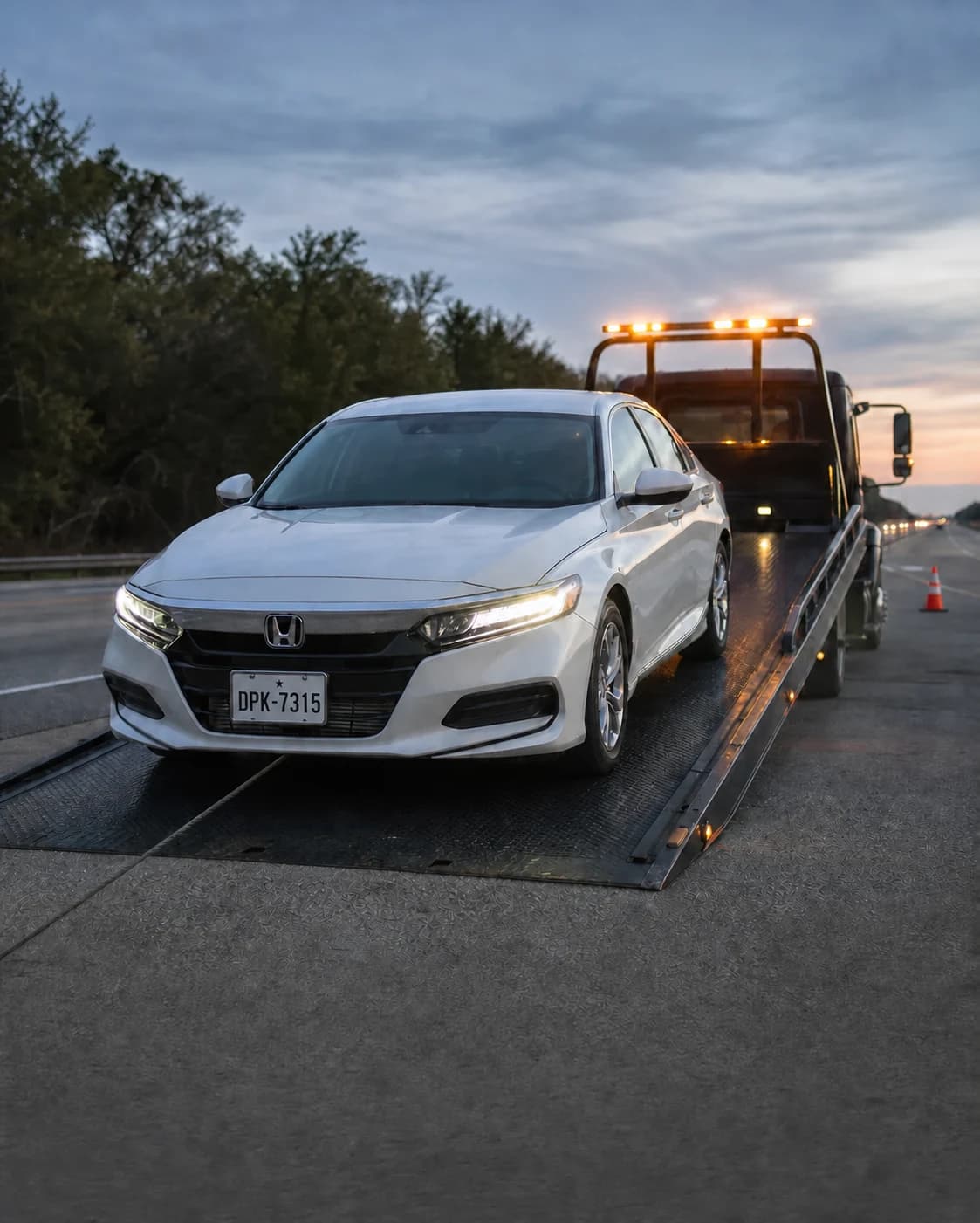 White sedan positioned on a flatbed tow truck beside a highway